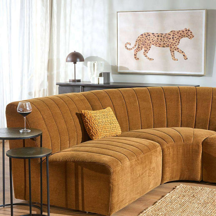 A curved sectional sofa upholstered in a warm camel-brown ribbed fabric, styled in a modern living room setting with a matching throw pillow, side tables, and wall art.