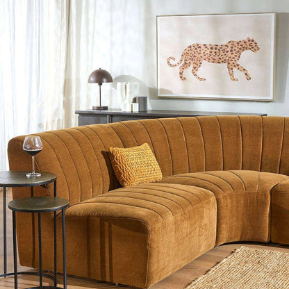 A curved sectional sofa upholstered in a warm camel-brown ribbed fabric, styled in a modern living room setting with a matching throw pillow, side tables, and wall art.