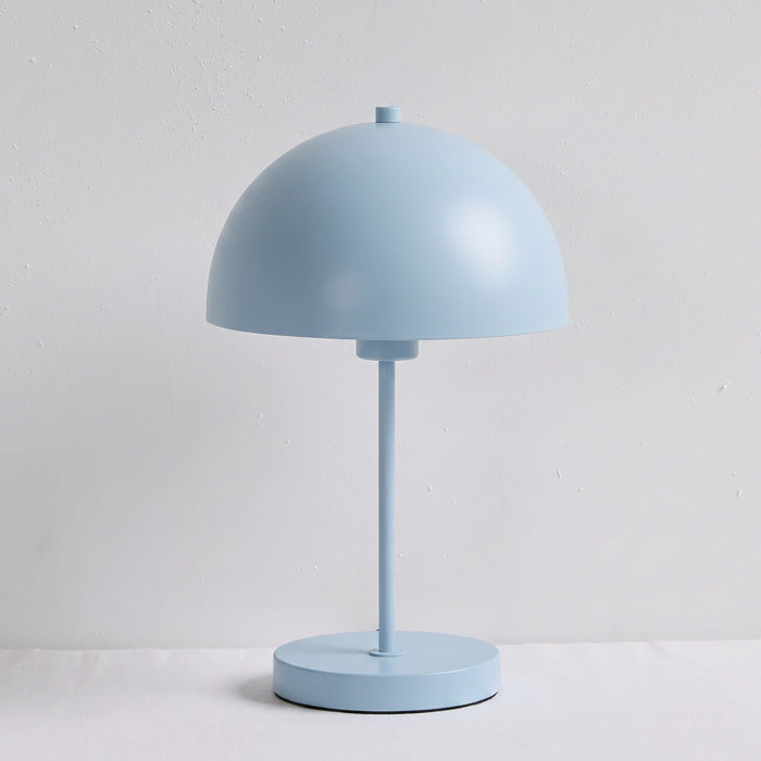 A light blue table lamp with a dome-shaped shade, a slender central stem, and a round base, photographed against a plain light gray wall and white surface.