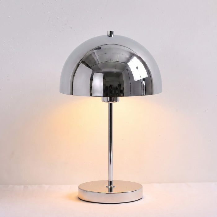 A modern table lamp with a polished chrome dome shade and circular base, illuminated with a warm yellow glow against a plain off-white background.