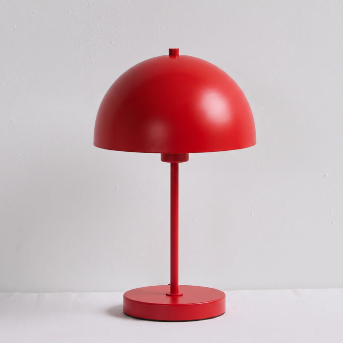A minimalist red table lamp with a dome-shaped shade, a slender stem, and a round base, set against a light gray textured wall and white surface.