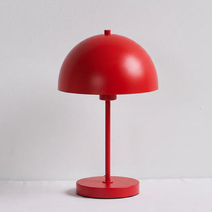 A minimalist red table lamp with a dome-shaped shade, a slender stem, and a round base, set against a light gray textured wall and white surface.