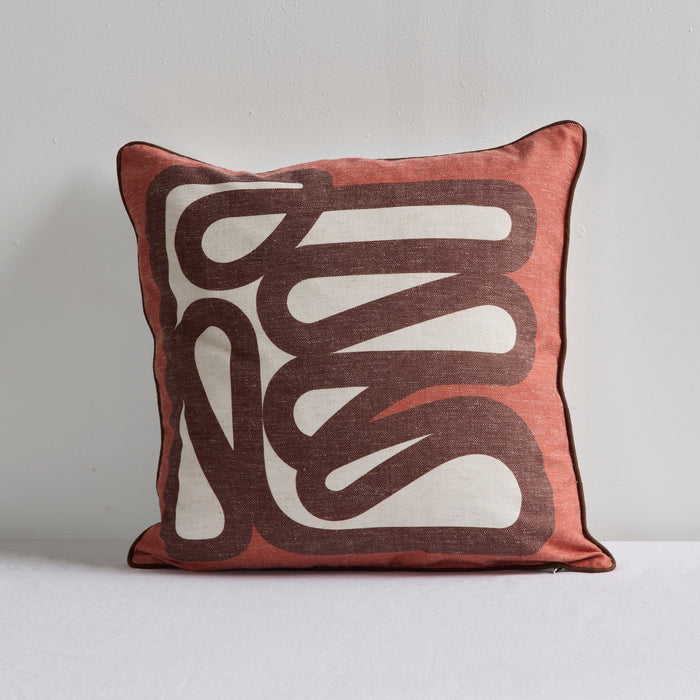 A square throw pillow with a terracotta pink border and a central cream and dark brown abstract wavy pattern, set against a neutral light gray background.