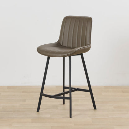 A modern counter height bar stool with a taupe-brown padded seat and ribbed backrest, supported by a black metal frame with splayed legs and a footrest, set against a neutral light gray wall and light wood floor.