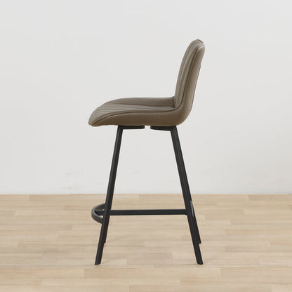 A modern counter stool with a taupe upholstered seat and curved backrest, supported by slim black metal legs, shown in profile against a white wall and light wood floor.