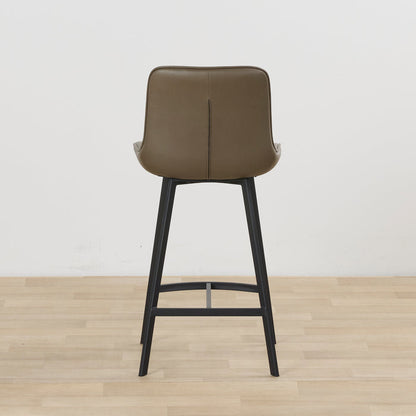 A modern bar stool with a taupe-brown upholstered seat and backrest on a black metal frame, set against a plain white wall and light wood floor.