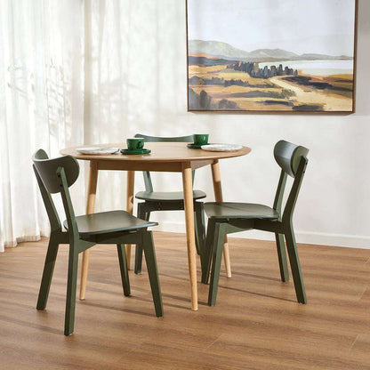 A round wooden dining table with four dark green chairs, styled with green cups and plates in a bright room with a wood floor and a landscape painting on the wall.