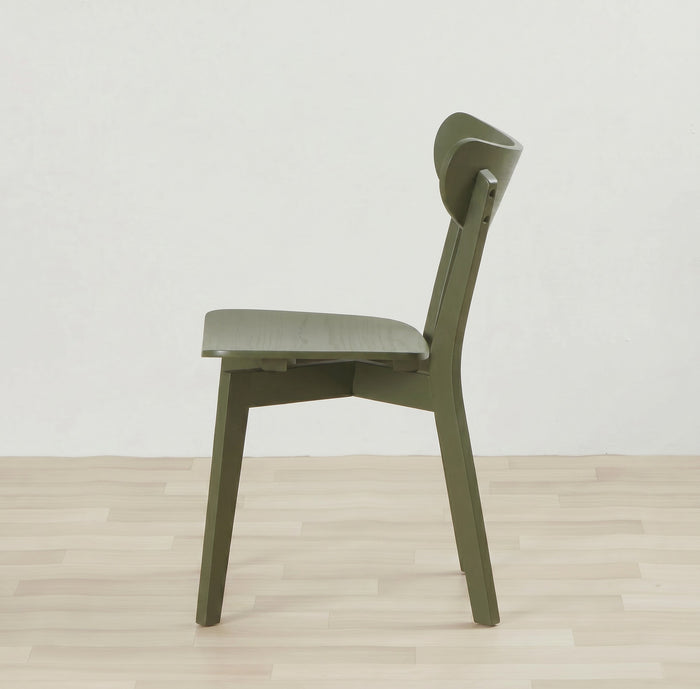 An olive green modern wooden dining chair shown from a side profile view, set against a minimalist off-white wall and light wood plank floor.