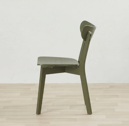 An olive green modern wooden dining chair shown from a side profile view, set against a minimalist off-white wall and light wood plank floor.